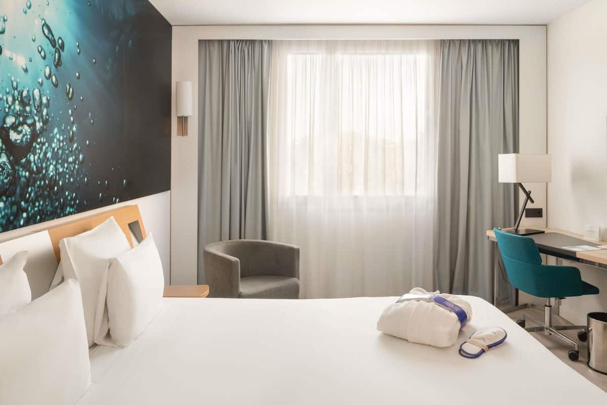 A hotel room with a bed, a desk, a chair, a lamp, and a painting on the wall.