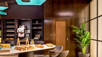 A modern kitchen with a coffee machine, shelves with jars, a wooden table with pastries, and a large potted plant.