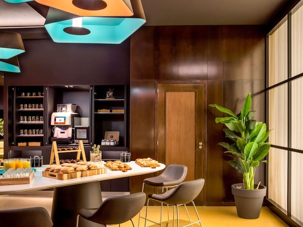 Una cocina moderna con una cafetera, estantes con frascos, una mesa de madera con pasteles y una planta grande en maceta.