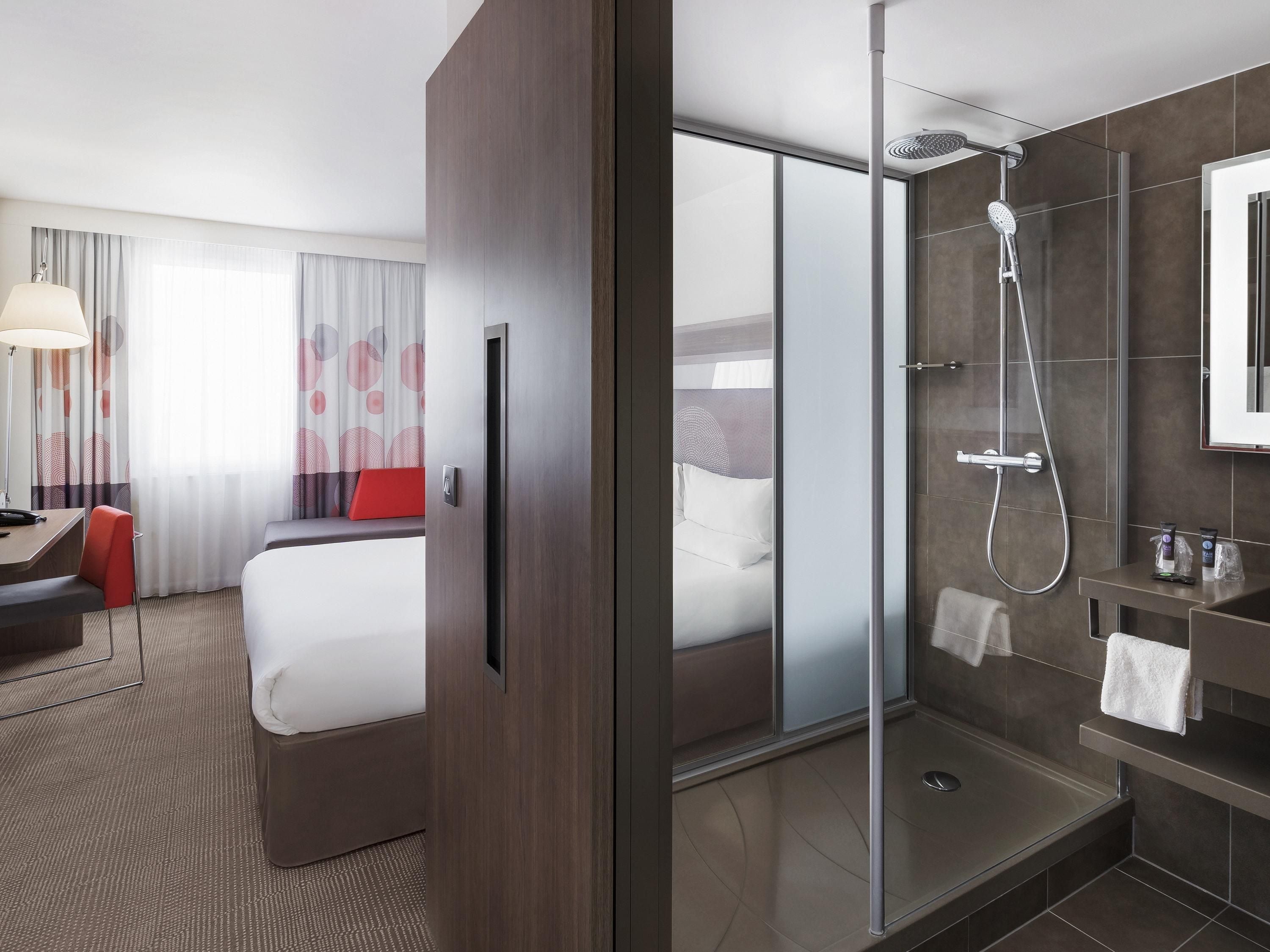 A modern hotel room with a bed, a desk, a chair, a shower, and a bathroom with a mirror and towel rack.