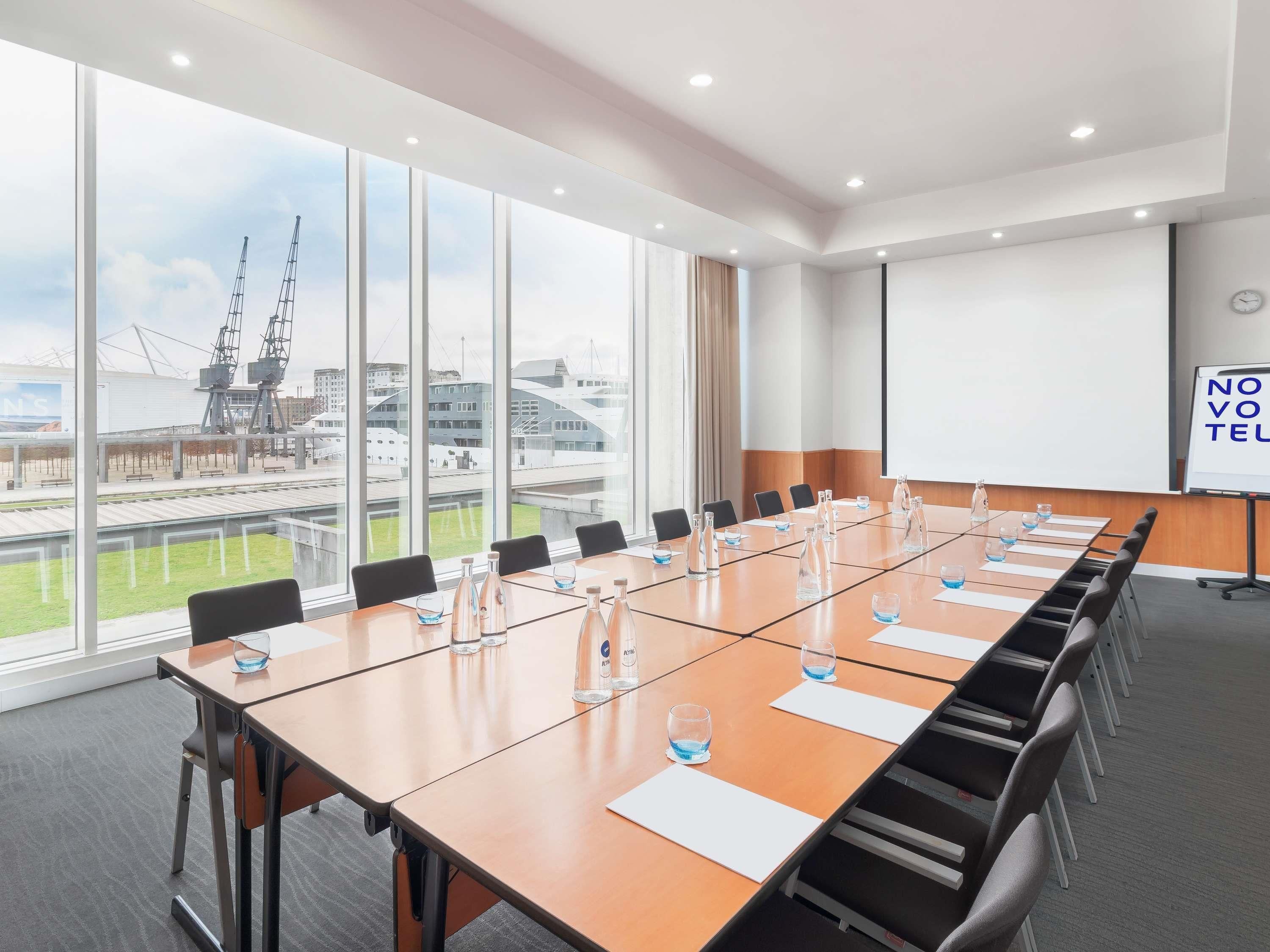 A conference room with a long table, chairs, a projector screen, and large windows with a view of a harbor.