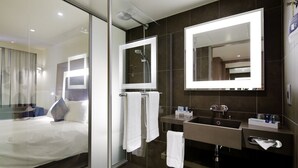 A modern hotel room with a large bed, a glass partition, a bathroom with a large mirror, a sink, and a shower. - Novotel London ExCeL (London)