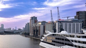 A waterfront hotel with modern architecture, a large white yacht docked nearby, and a city skyline in the background. - Novotel London ExCeL (London)
