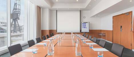 A conference room with a long table, chairs, a projector screen, and a view of a construction crane outside.