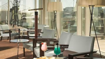 Ein stilvoller Lounge-Bereich mit bequemen Sitzgelegenheiten, einem Glastisch mit Snacks und Getränken sowie großen Fenstern mit Blick auf die Stadtlandschaft.