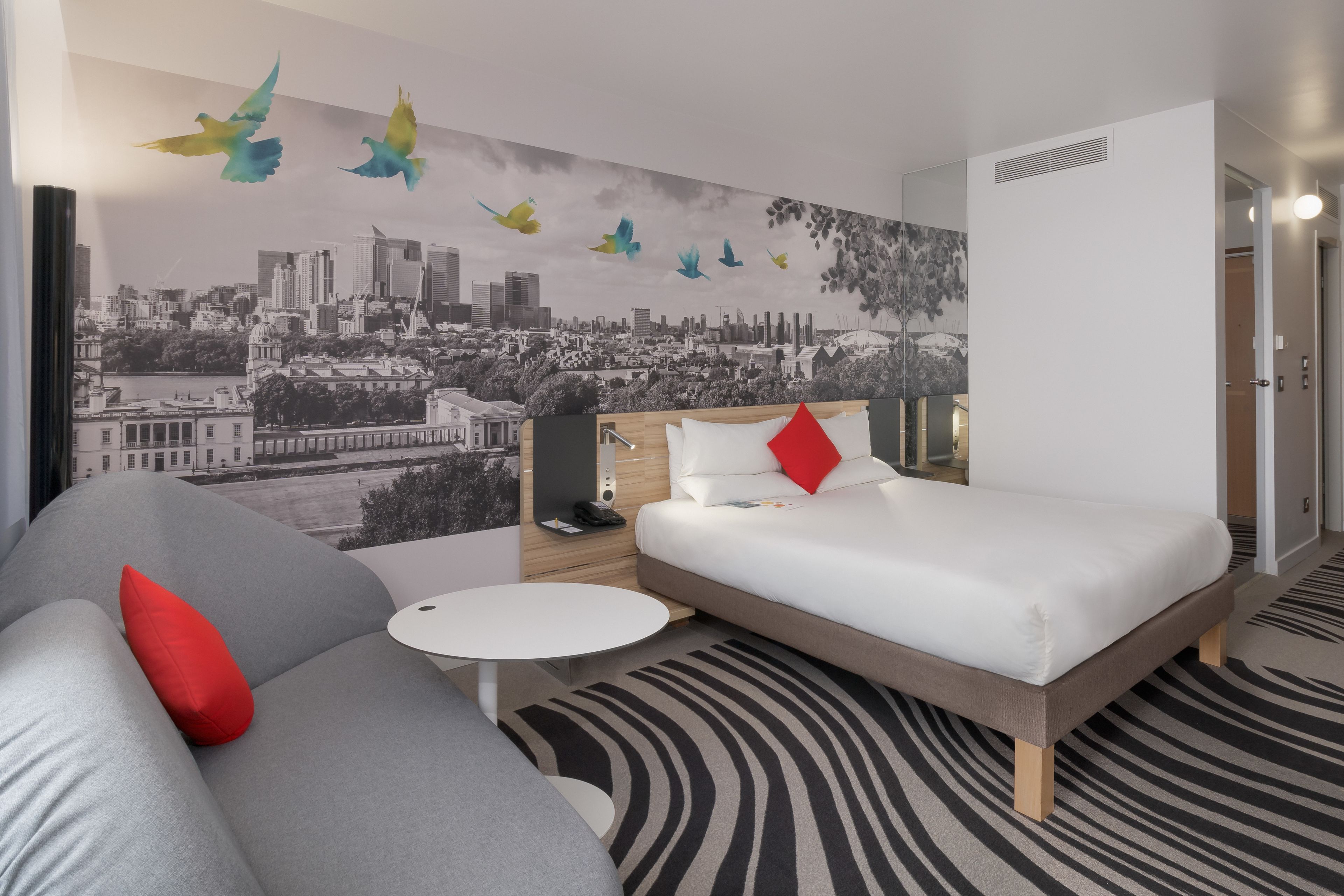 A modern hotel room with a cityscape mural, a bed with white bedding and red pillows, a gray sofa with a red pillow, a small white table, and a patterned rug.