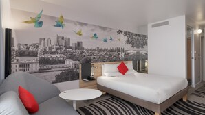 A modern hotel room with a cityscape mural, a bed with white bedding and red pillows, a gray sofa with a red pillow, a small white table, and a patterned rug.
