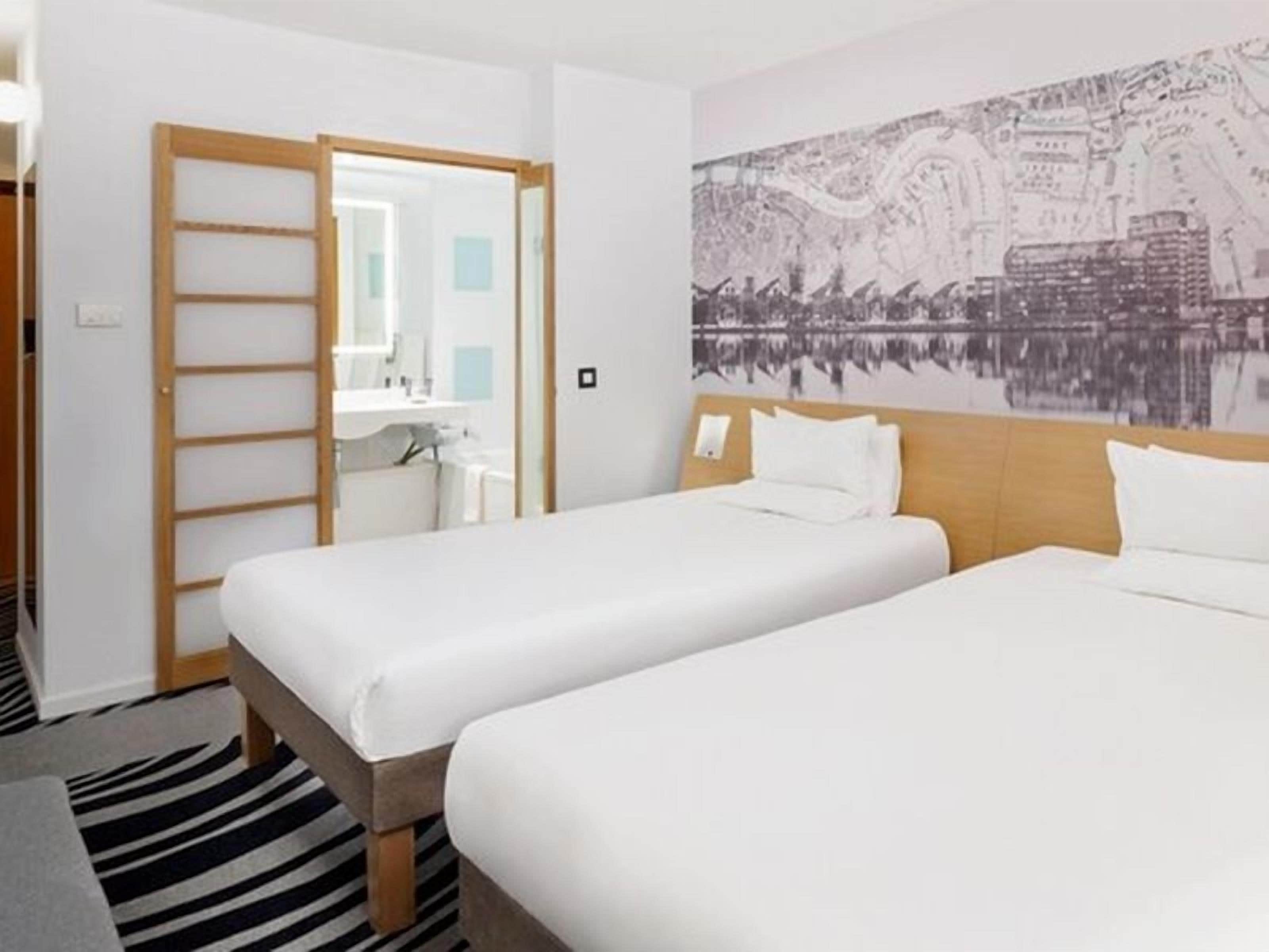 A hotel room with two beds, a wooden headboard, a cityscape mural, a bathroom with a sink, and a striped rug.