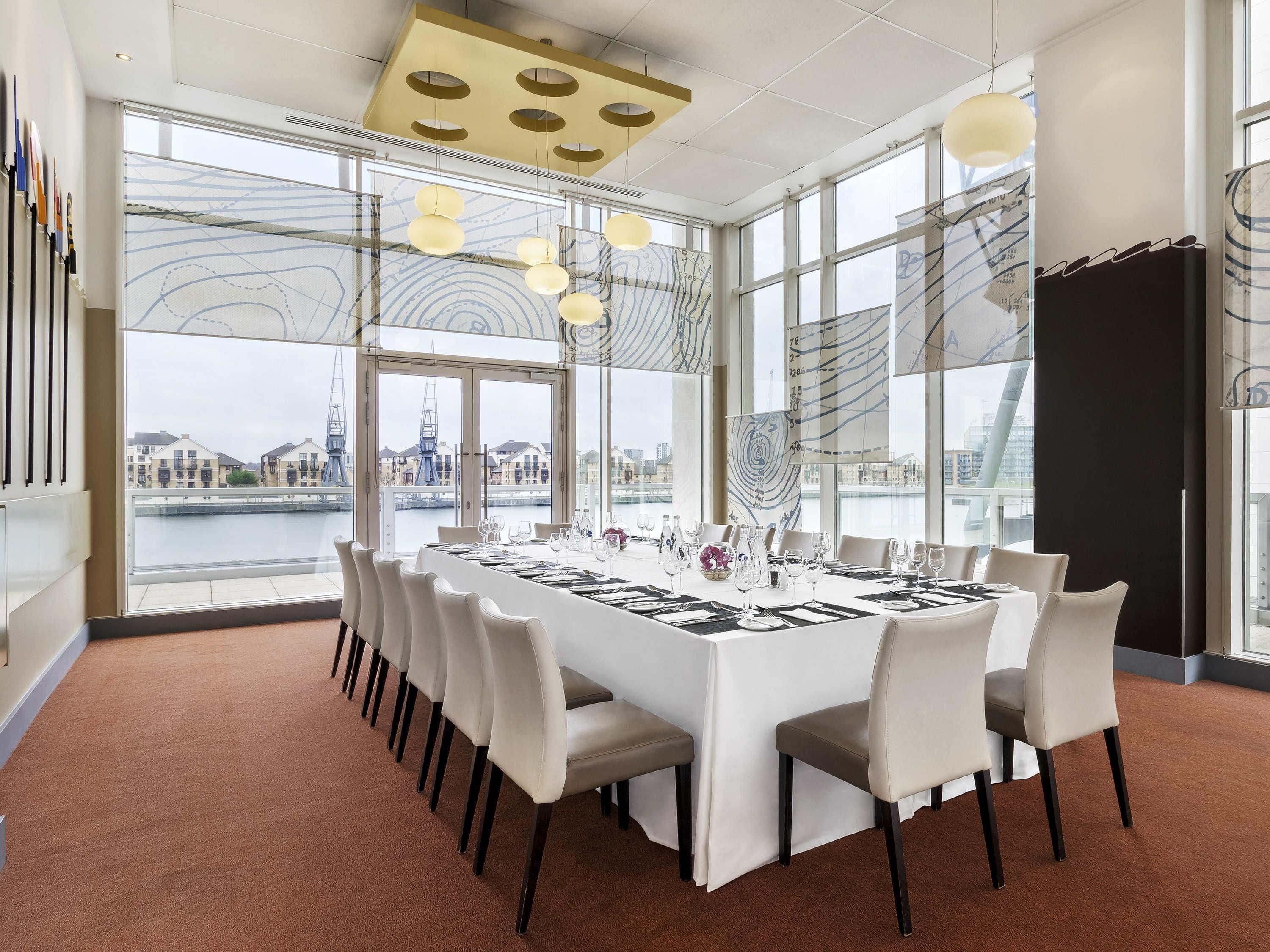 A conference room with a large table set for a meeting, large windows with a view of a river and buildings, and modern lighting fixtures.