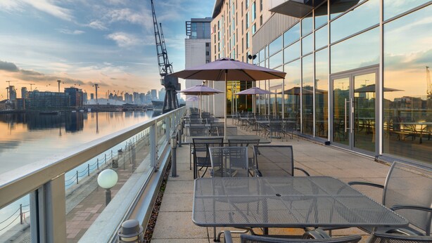 A rooftop terrace with tables and chairs, overlooking a river with a city skyline in the background. - Novotel London ExCeL (London)