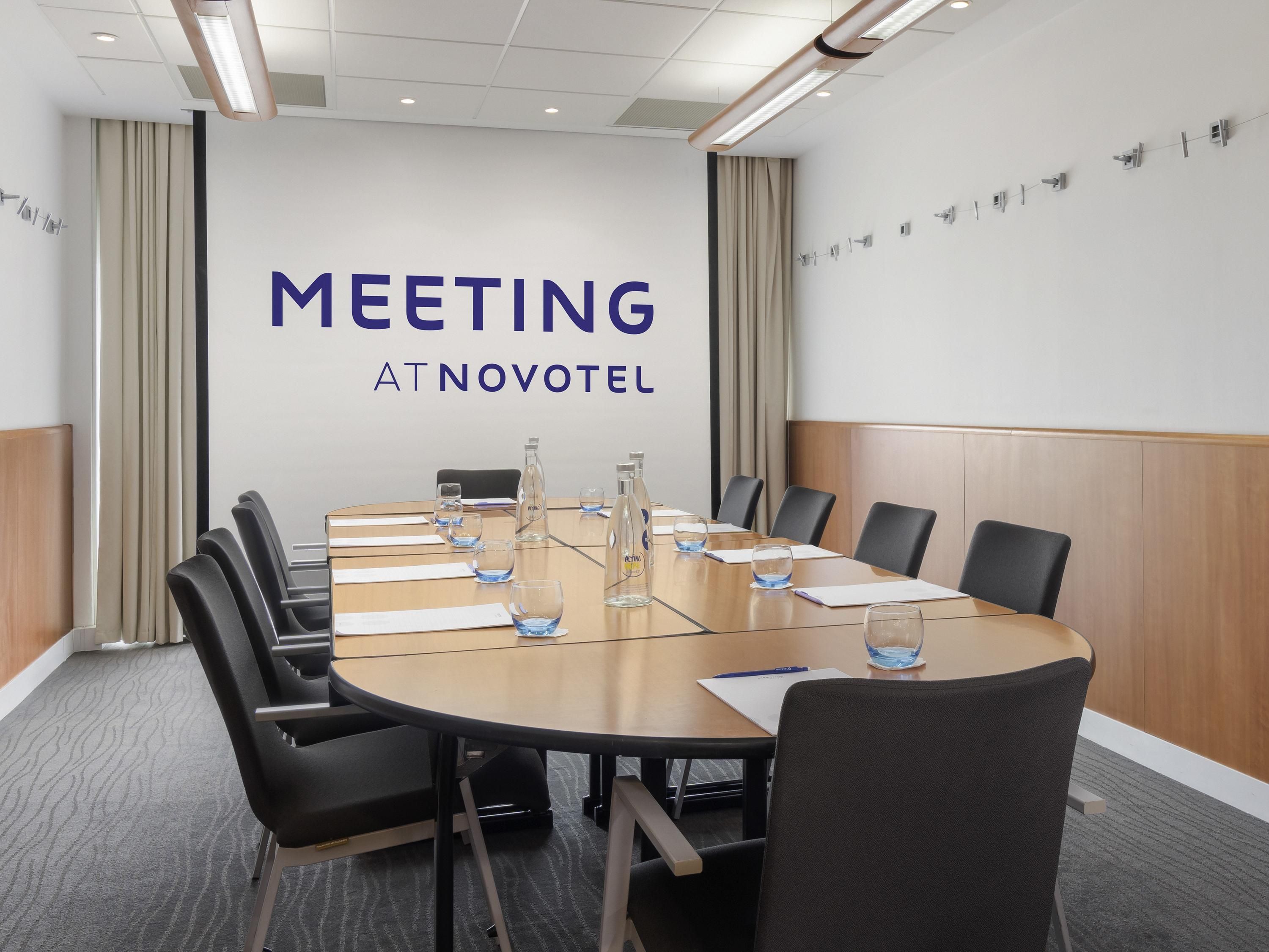 A meeting room with a large table, chairs, and a wall with 'MEETING AT NOVOTEL' text.