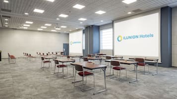 Un salón de conferencias con mesas y sillas, una pantalla de proyector mostrando el logotipo de ILUNION Hotels y un amplio ventanal.