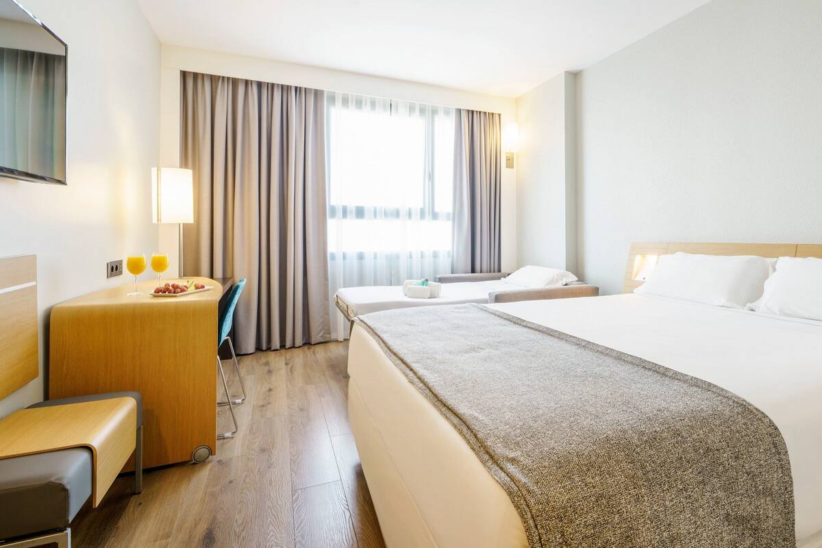 A hotel room with a large bed, a desk, a chair, a TV, and a window with curtains.