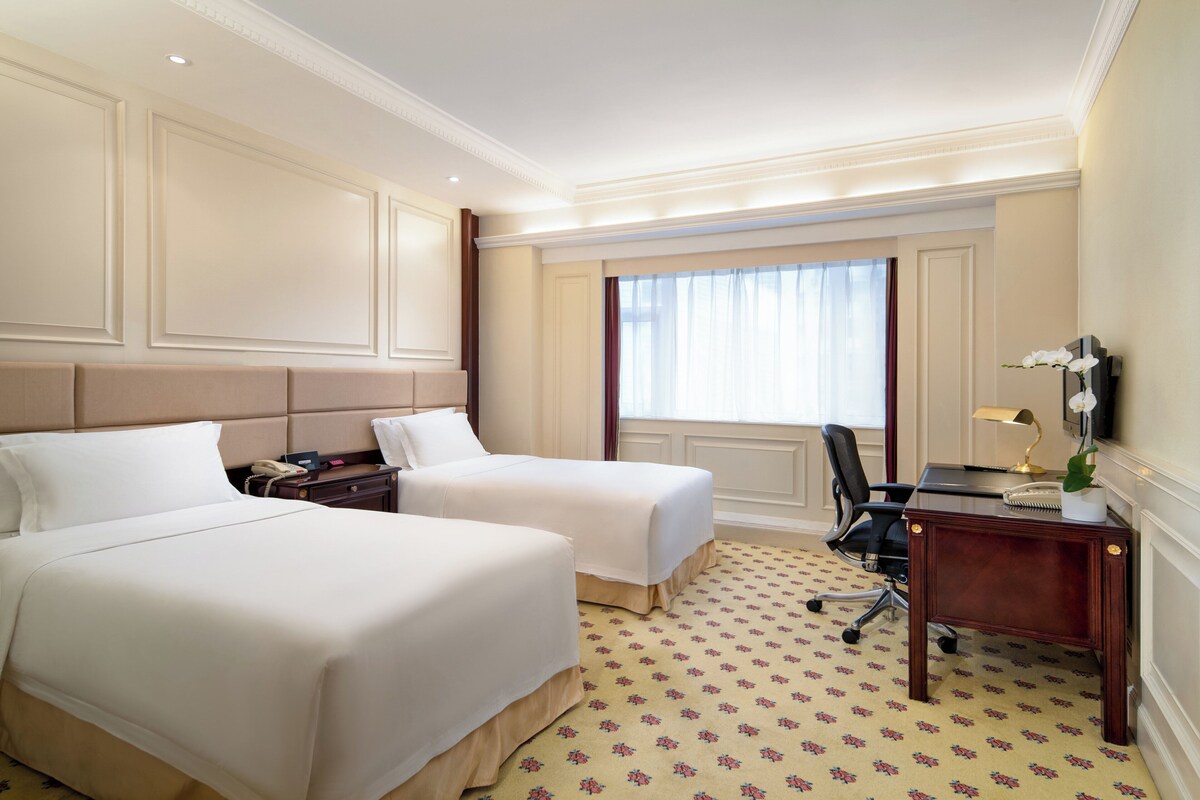 Une chambre d’hôtel avec deux lits, un bureau, une chaise, un téléphone et une fenêtre avec des rideaux.