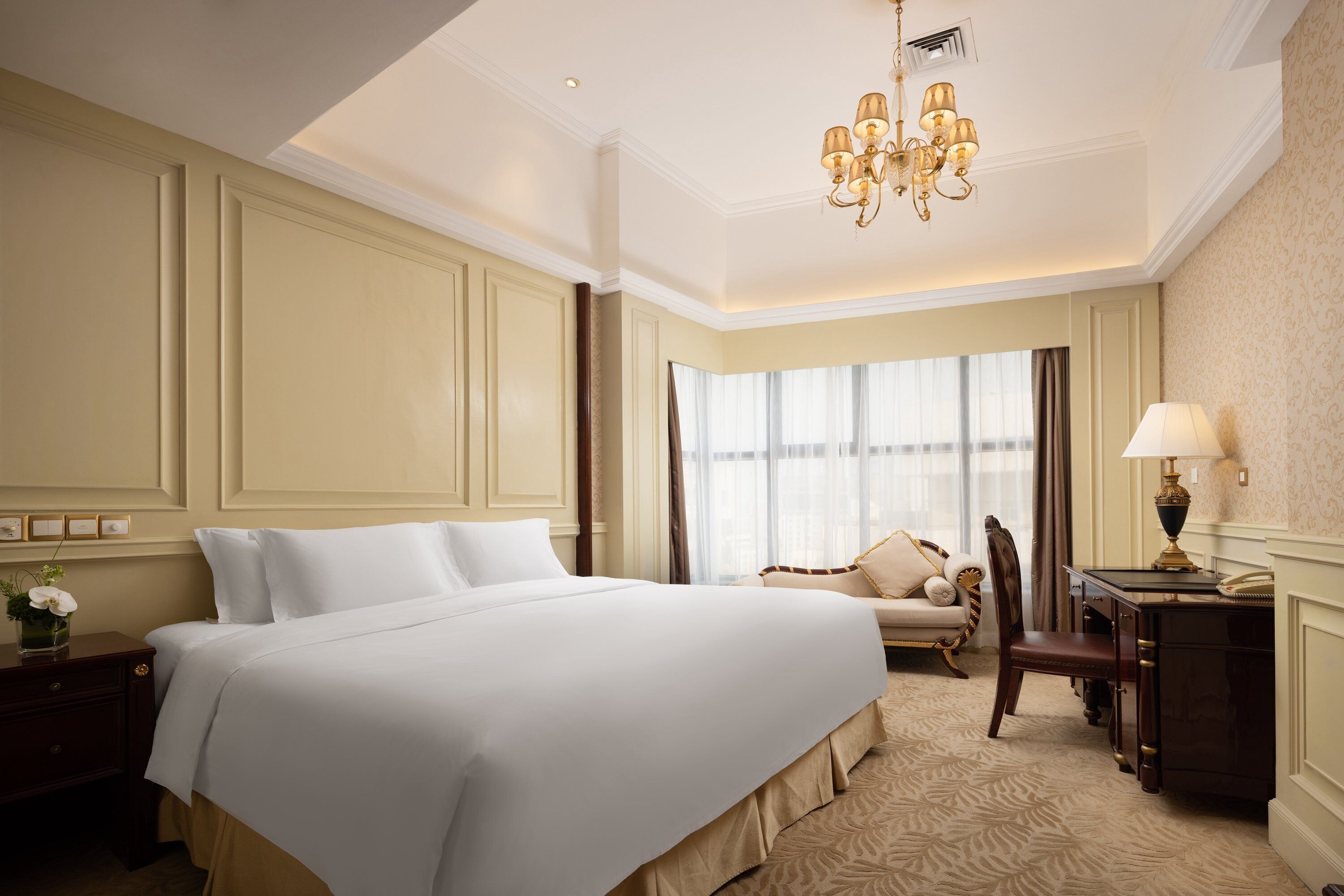 A hotel room with a large bed, a desk, a chair, a lamp, and a chandelier.