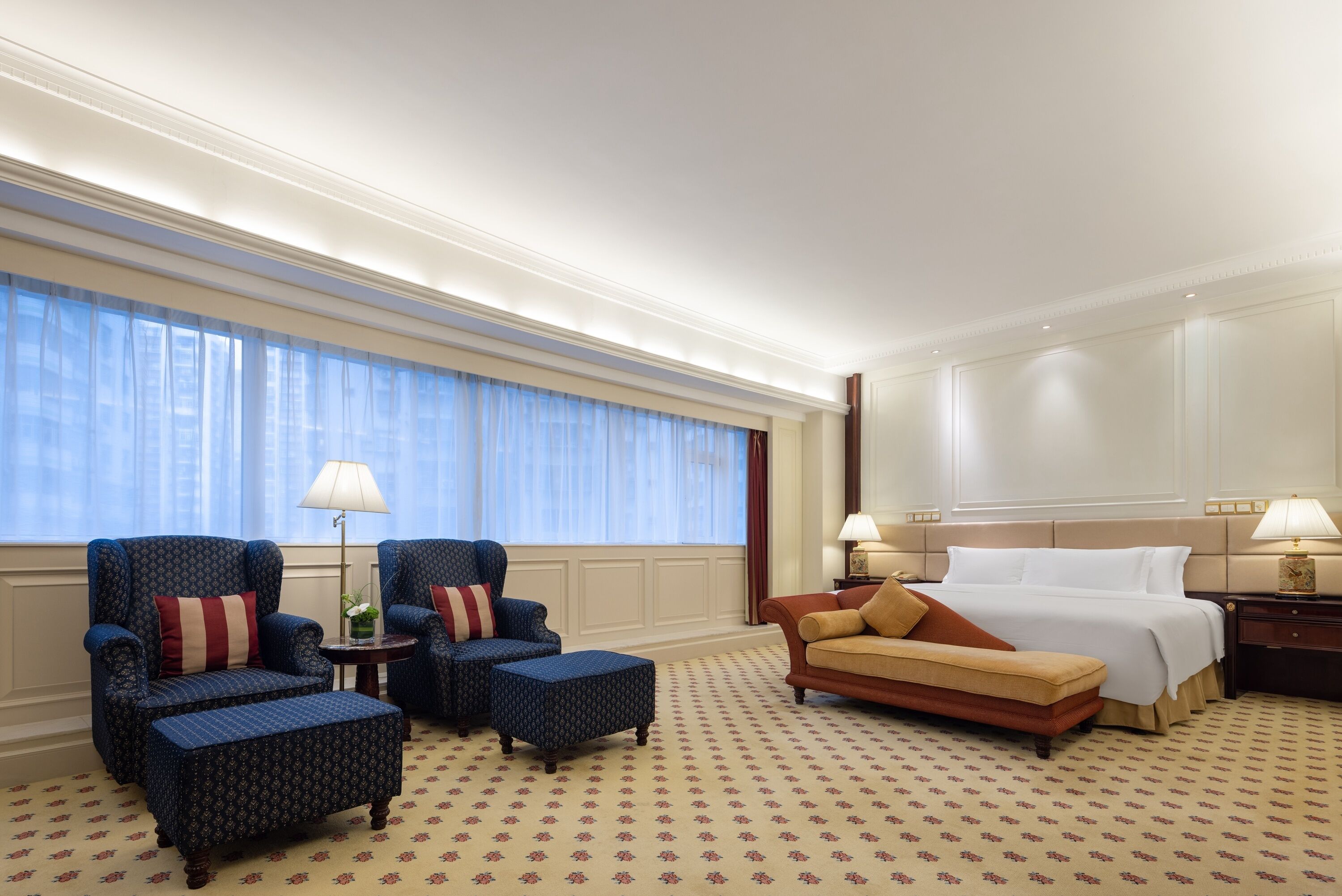 Quarto de hotel com uma cama grande, duas poltronas, um sofá e uma mesinha de cabeceira com luminárias.