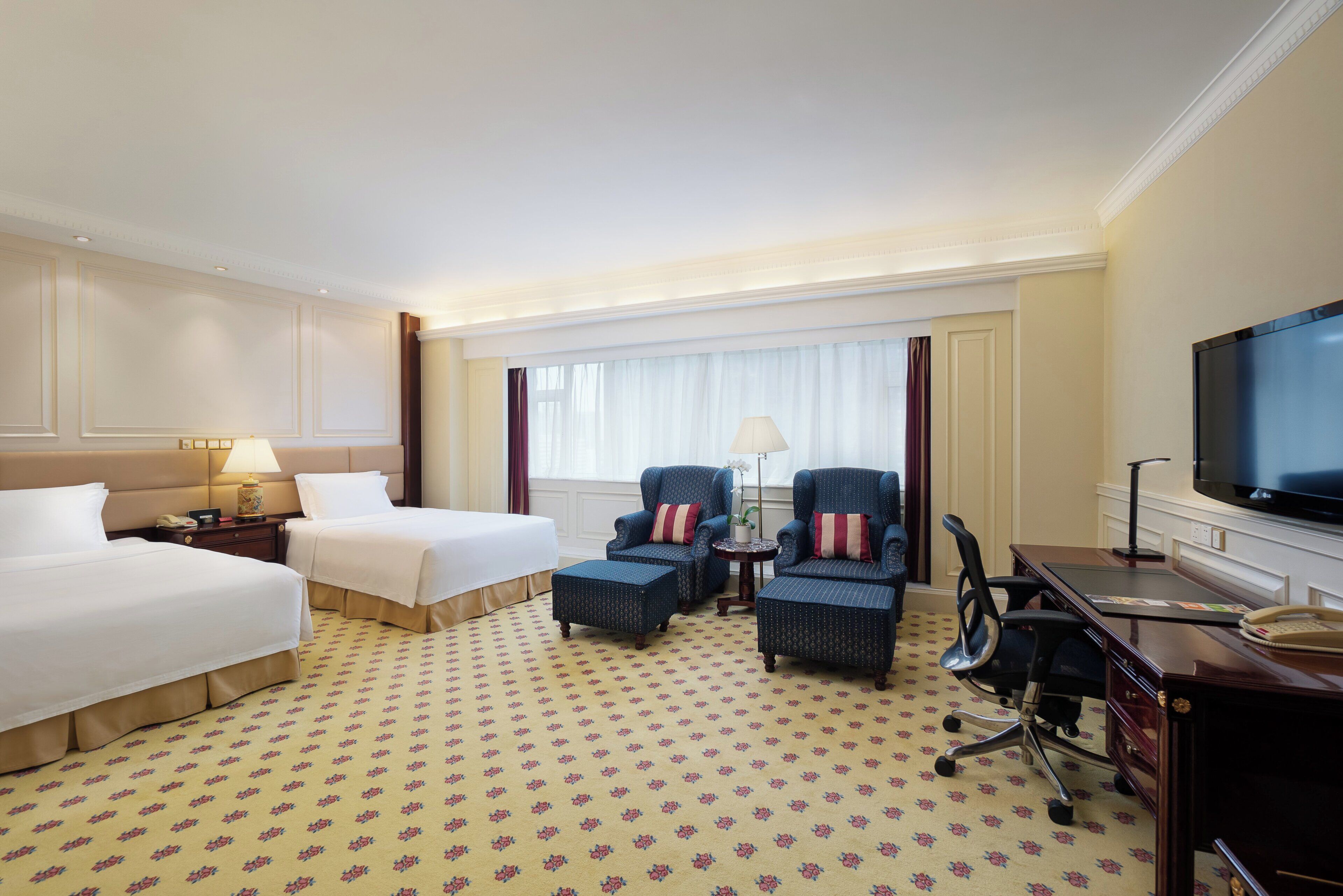 A hotel room with two beds, a desk, a television, and a window with curtains.