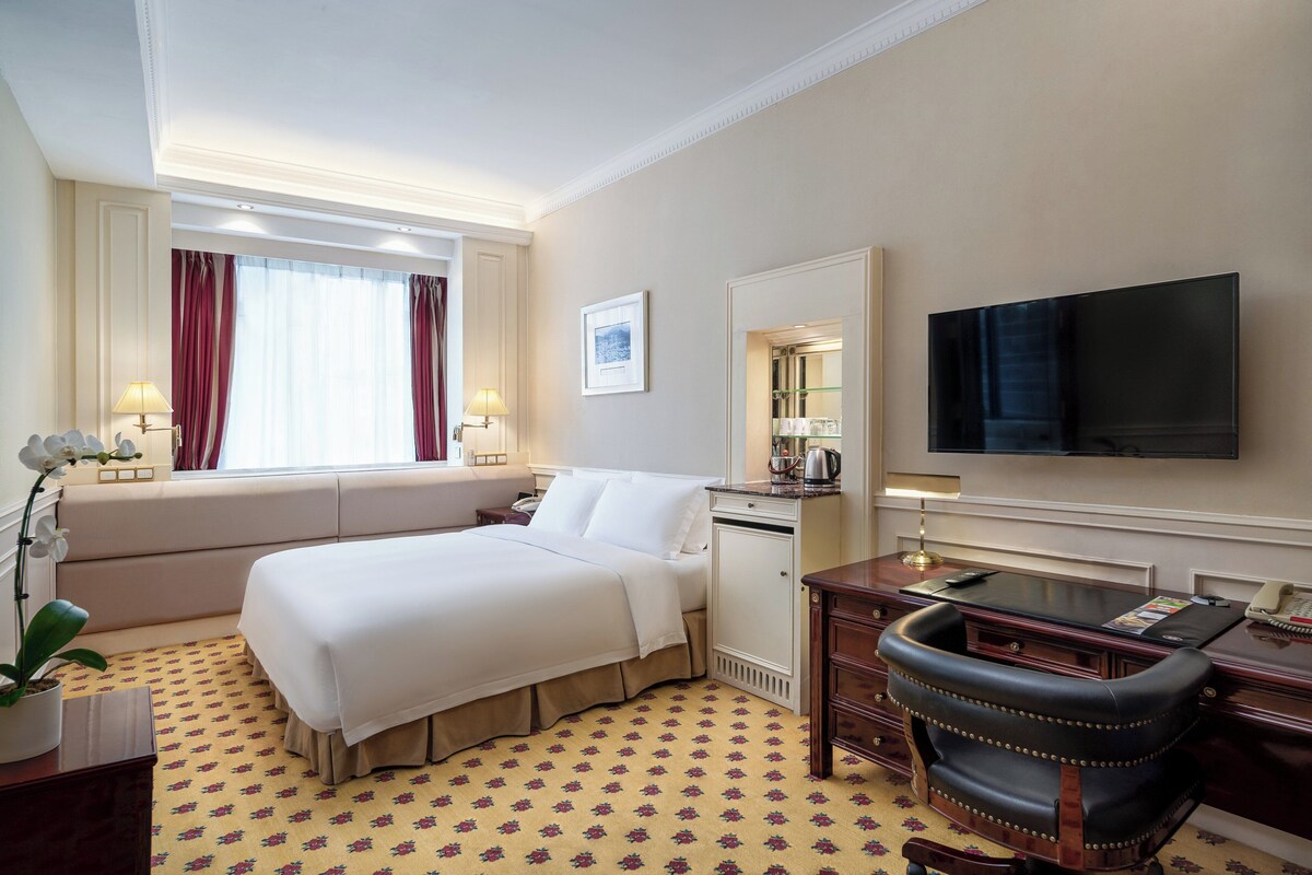 Une chambre d’hôtel avec un grand lit, un bureau avec une chaise, une télévision et une fenêtre avec des rideaux.