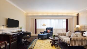 A hotel room with a flat-screen TV, a desk with a telephone, a blue armchair, a striped sofa, and a patterned rug.