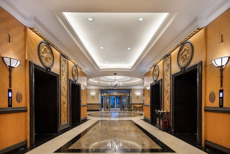 A hotel lobby with a polished marble floor, ornate wall decorations, and a central chandelier.