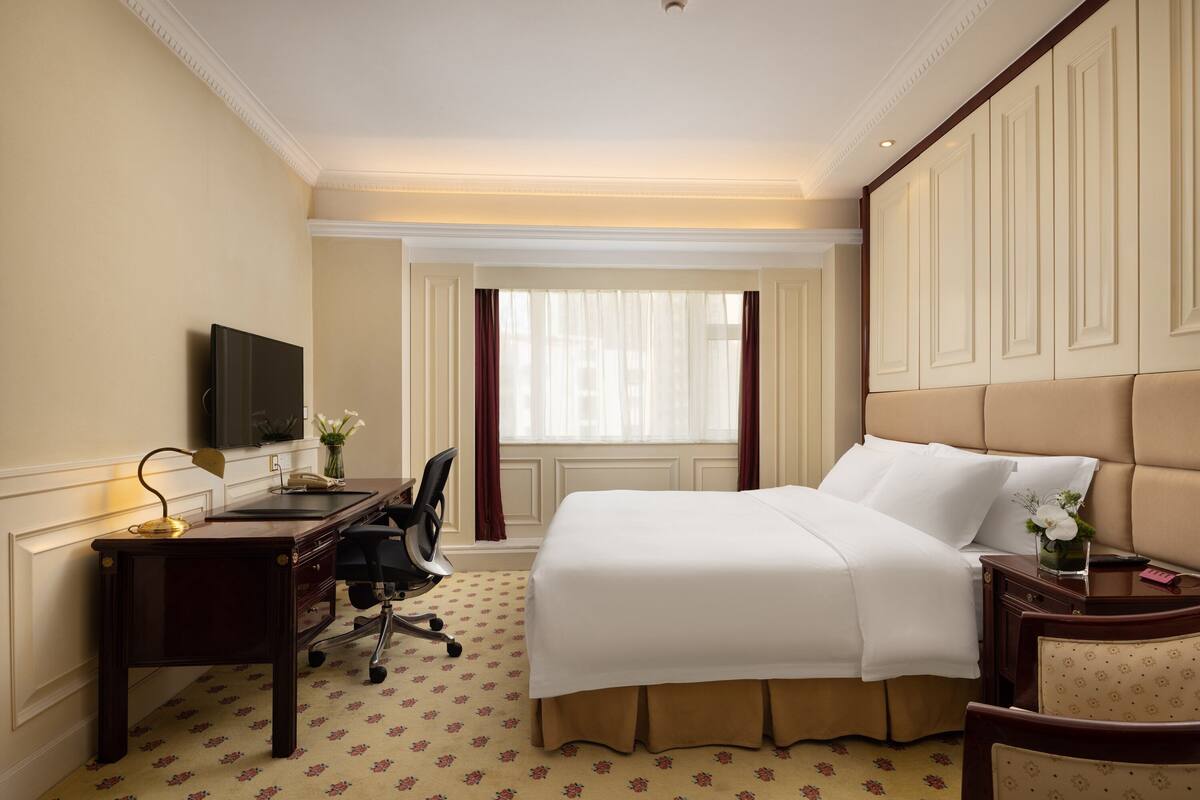 Ein Hotelzimmer mit einem großen Bett, einem Schreibtisch mit Stuhl, einem Fernseher und einem Fenster mit Vorhängen.
