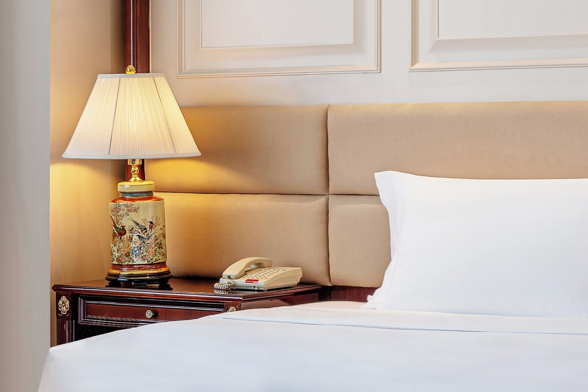 Un lit bien fait, avec un oreiller blanc et une tête de lit beige. Une table de chevet porte une lampe avec un pied décoratif et un téléphone.