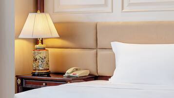 A neatly made bed with a white pillow and a beige headboard. A bedside table holds a lamp with a decorative base and a telephone.