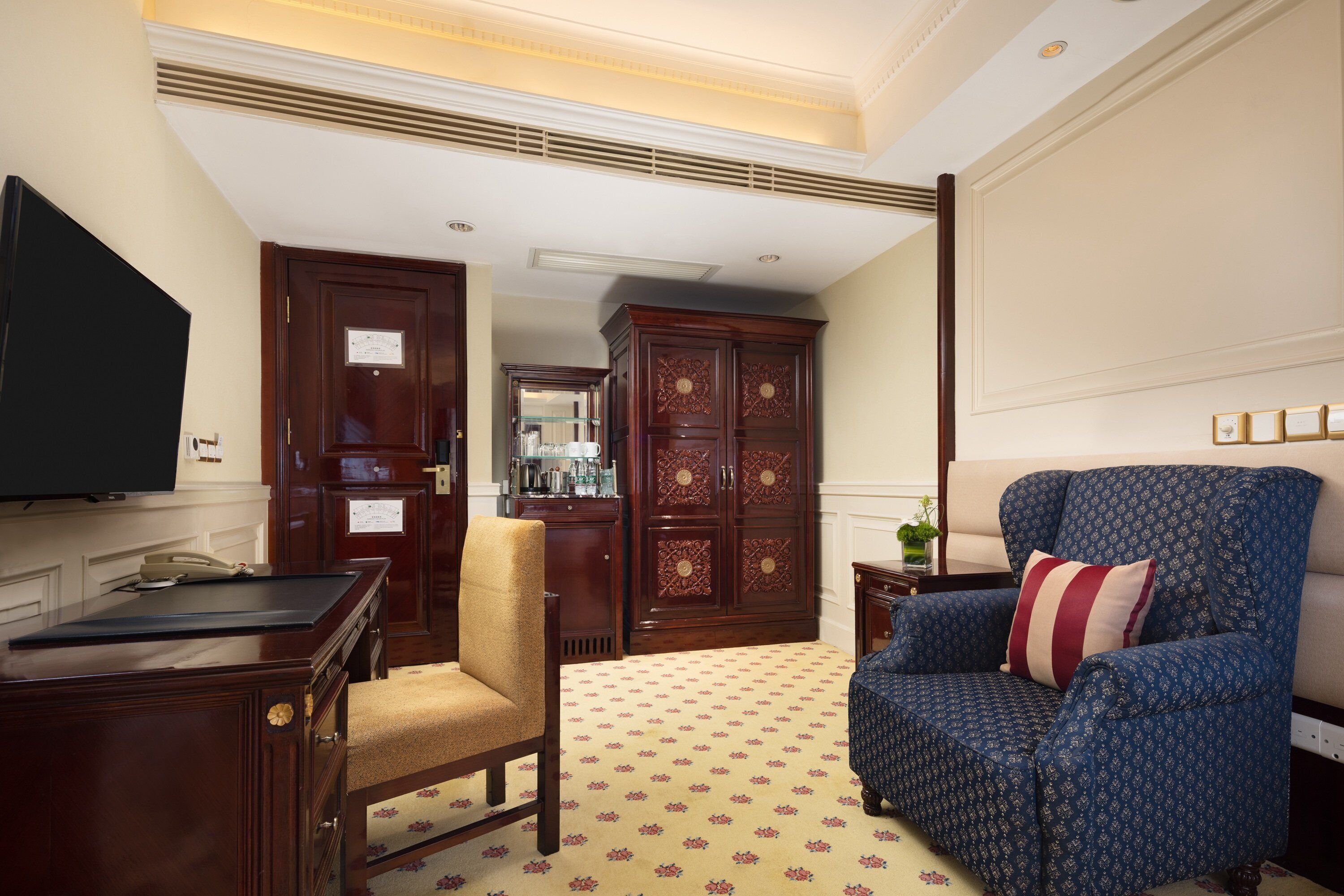 Une chambre d’hôtel avec un grand bureau en bois, une télévision, un fauteuil bleu avec un coussin à rayures et une moquette à motifs.