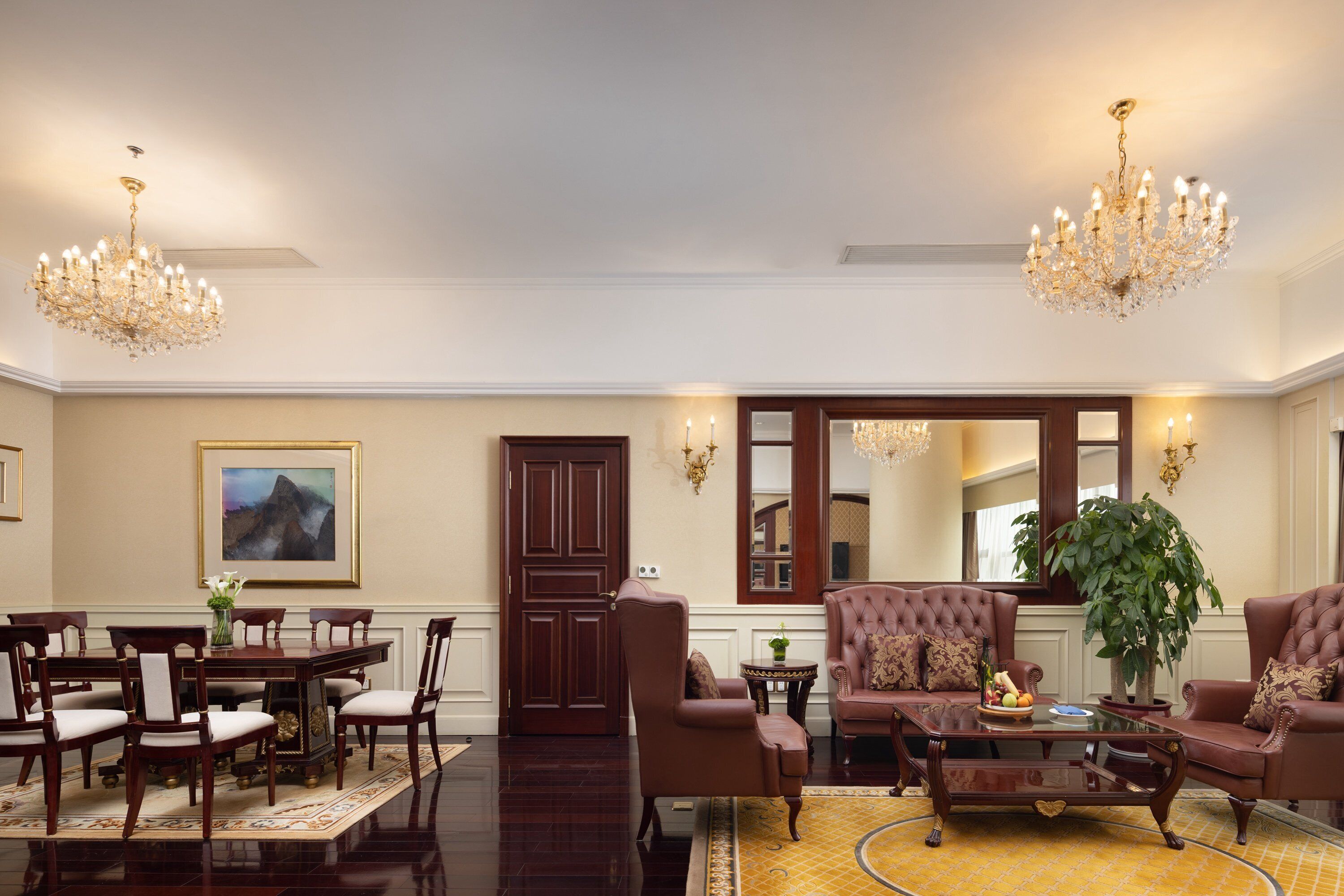 A formal living room with a dining area, featuring a large painting, a grand chandelier, and a large potted plant.