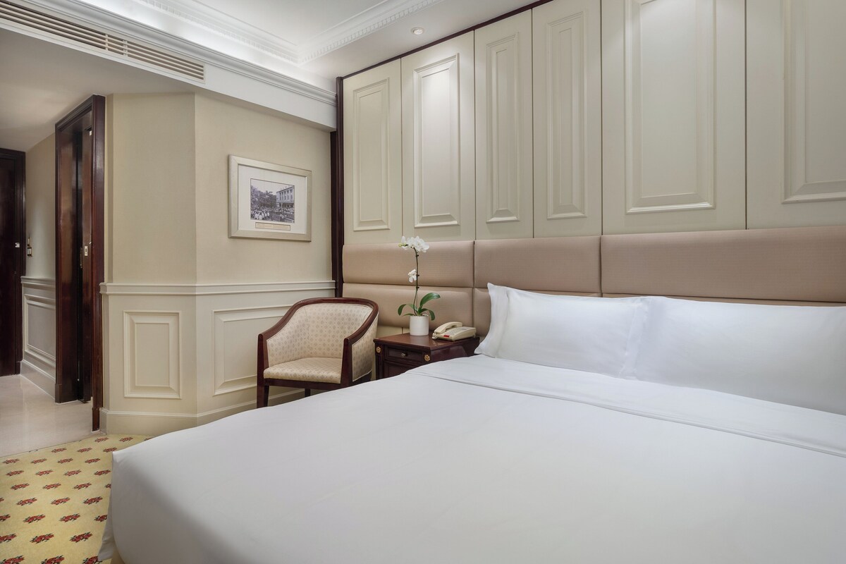 Une chambre d’hôtel avec un grand lit, une chaise, une table de chevet avec un téléphone et un vase contenant une fleur, ainsi qu’un tableau encadré au mur.