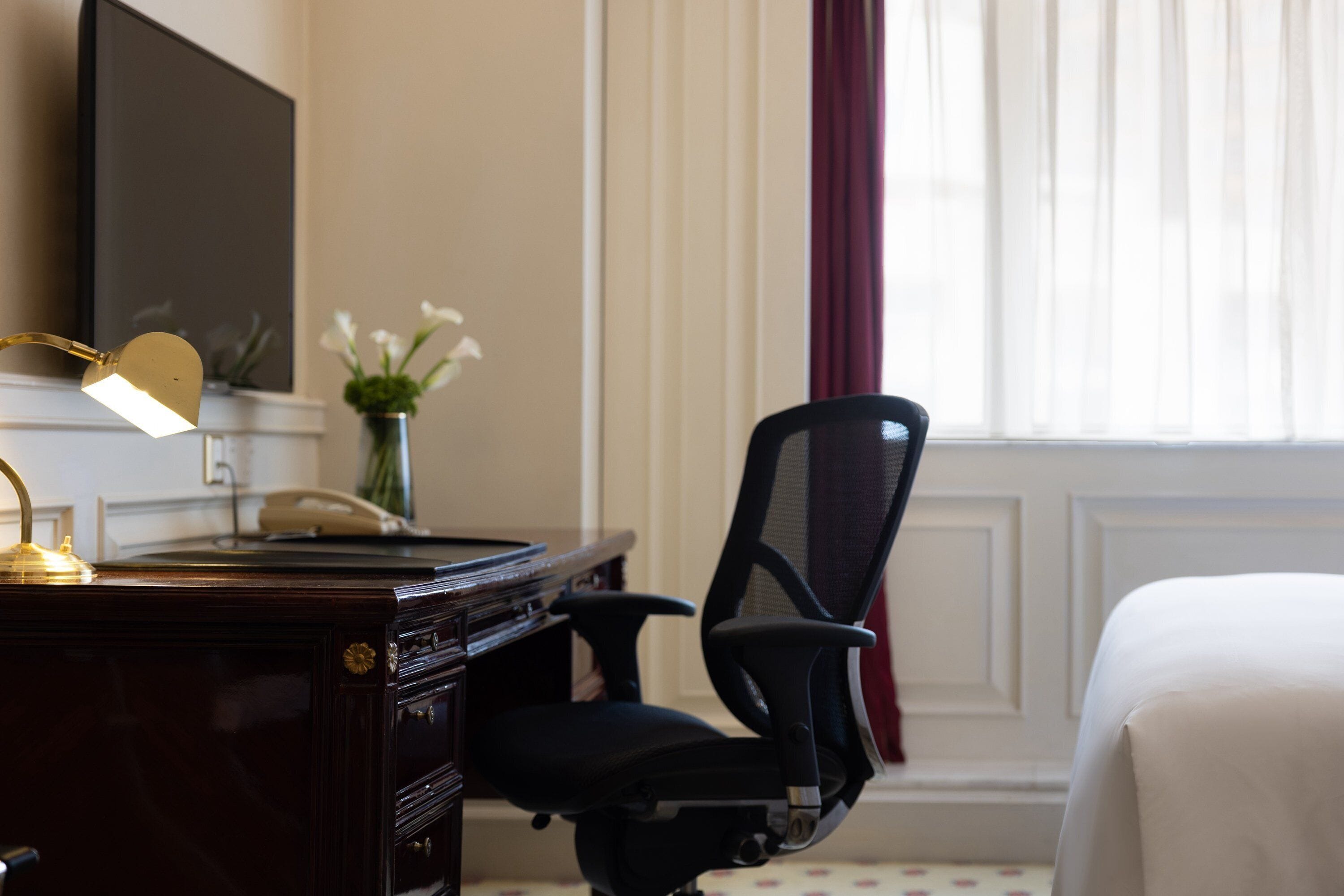 Quarto de hotel com escrivaninha, cadeira, cama, televisão na parede e janela com cortinas.