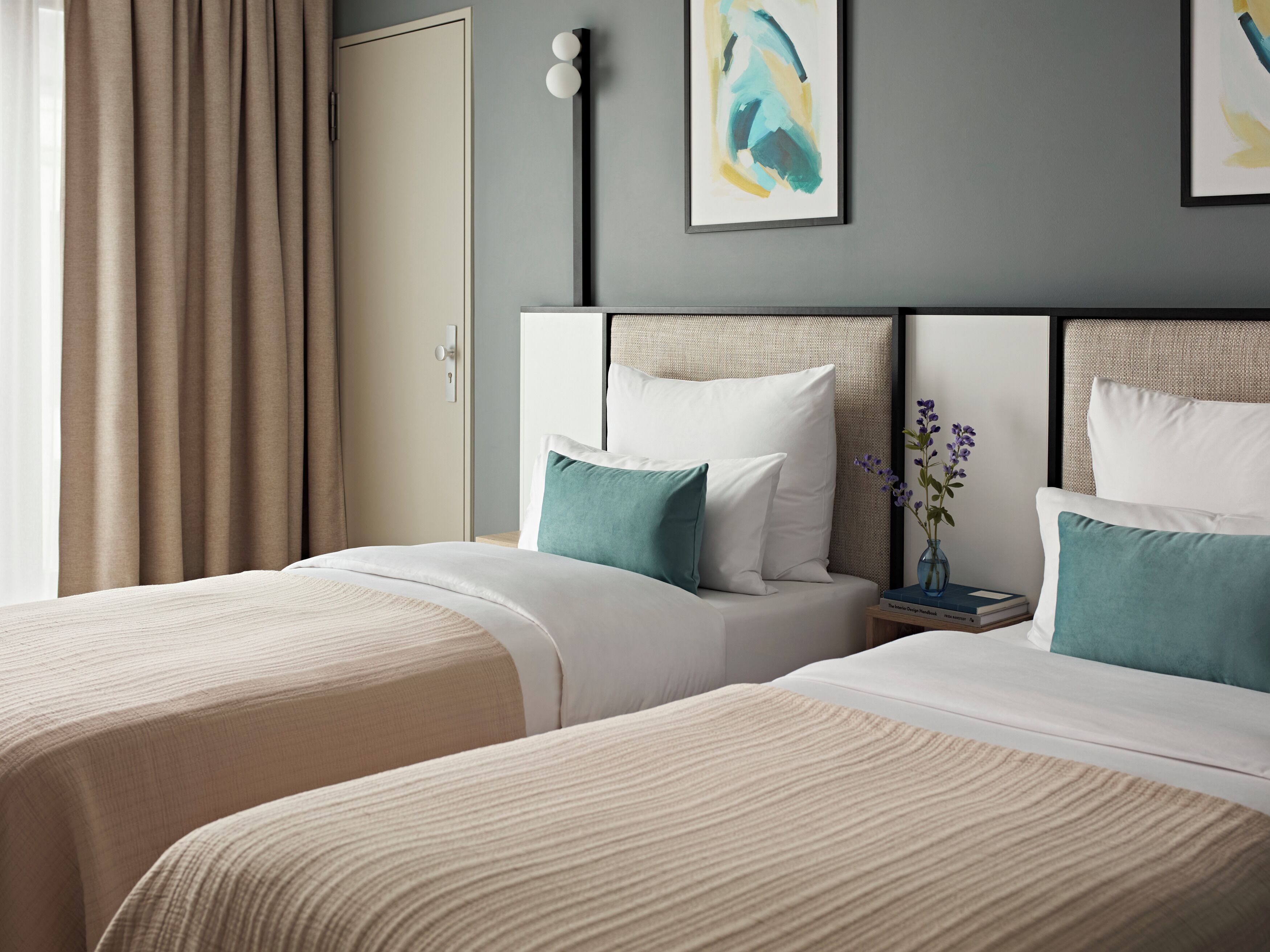Une chambre d’hôtel avec deux lits, une literie de couleur beige, une table de chevet ornée d’un vase de fleurs et un tableau accroché au mur.