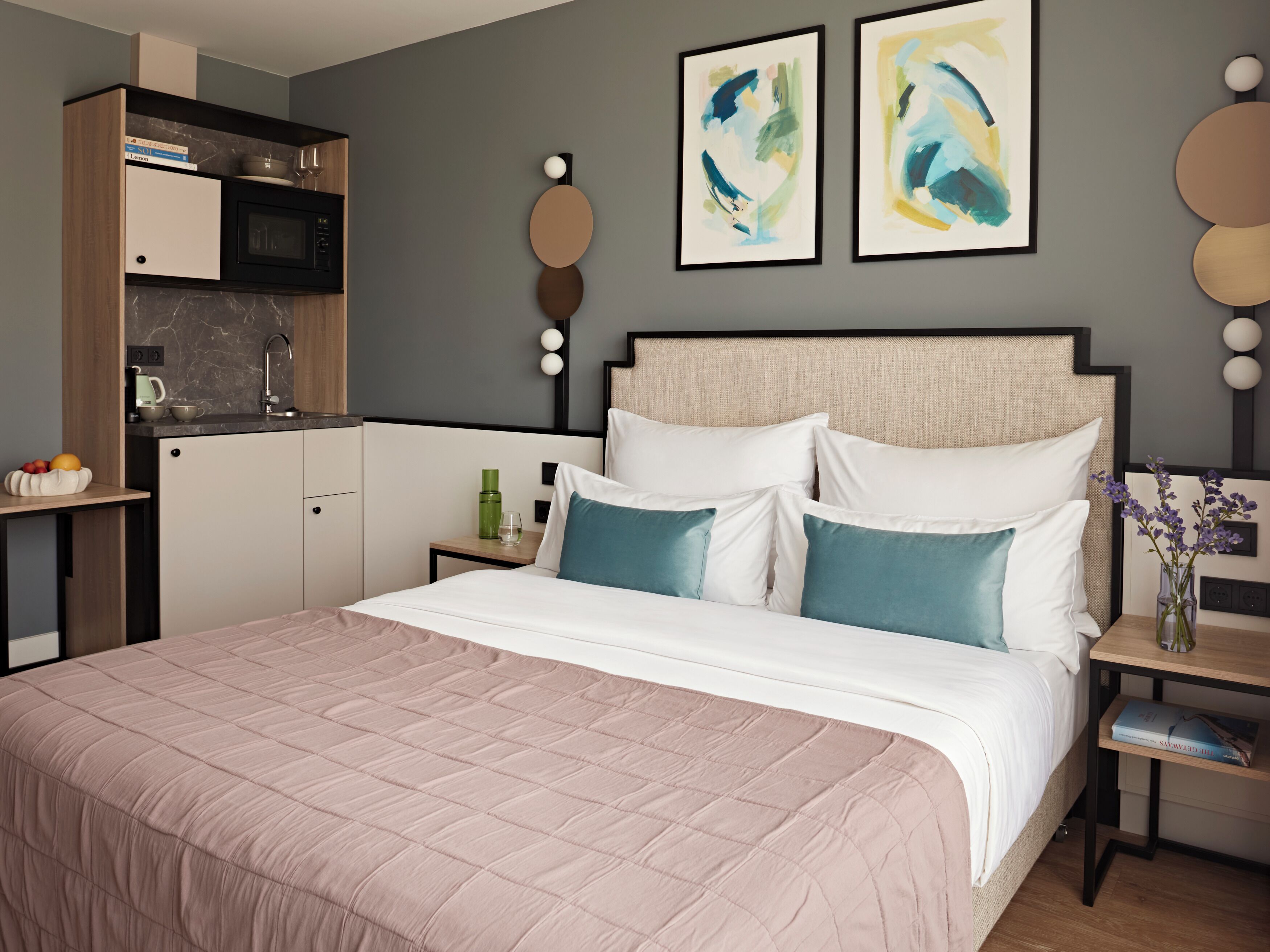 Une chambre à coucher moderne avec un grand lit, une table de chevet, une commode, un coin cuisine intégré et une œuvre d’art abstraite au mur.