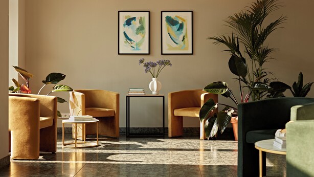A modern lobby with two framed abstract paintings, a variety of seating options including a sofa and chairs, a small table with a vase of flowers, and several potted plants.