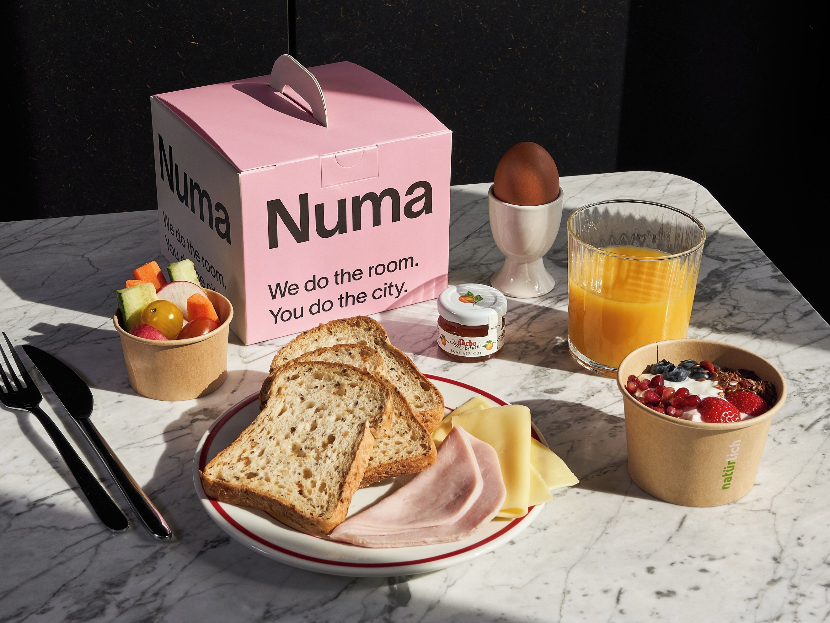 A breakfast setup with a pink box labeled 'Numa,' a bowl of fruit, a glass of orange juice, a plate with bread, cheese, and ham, and a cup of yogurt with berries.