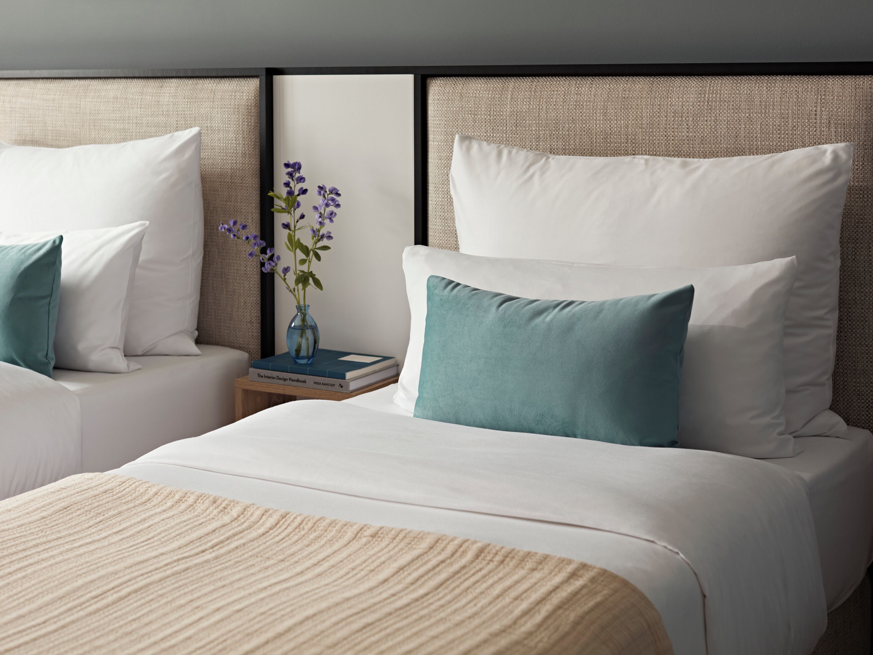 A hotel room with two beds, each with white linens and teal pillows. There is a small bedside table with a book and a vase with flowers.