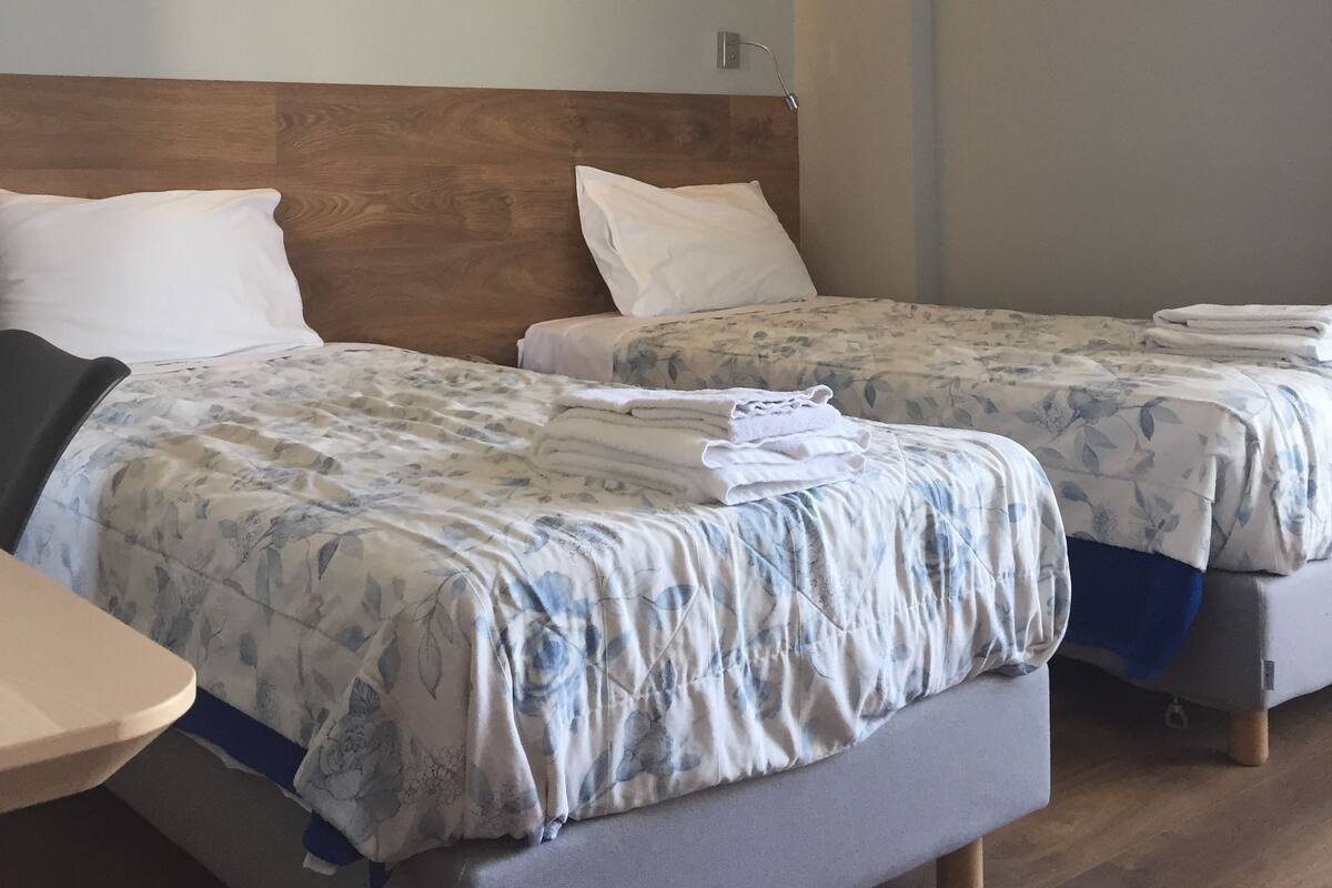 A hotel room with two beds, wooden headboards, a bedside table, and an air conditioning unit.