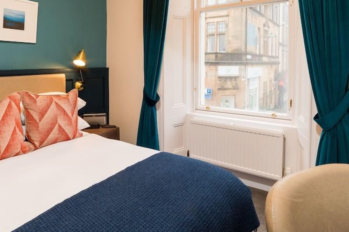 A hotel room with a bed, a chair, a nightstand with a lamp, a window with curtains, and a view of buildings outside.