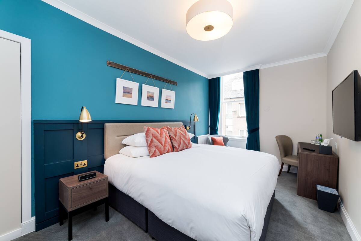 A hotel room with a large bed, a bedside table, a desk with a chair, a TV, and framed pictures on the wall.
