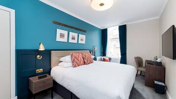 A hotel room with a large bed, a bedside table, a desk with a chair, a TV, and framed pictures on the wall.