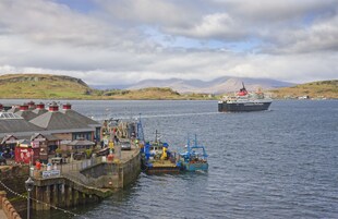 Un puerto con un barco grande, un muelle con edificios y un paisaje montañoso al fondo.