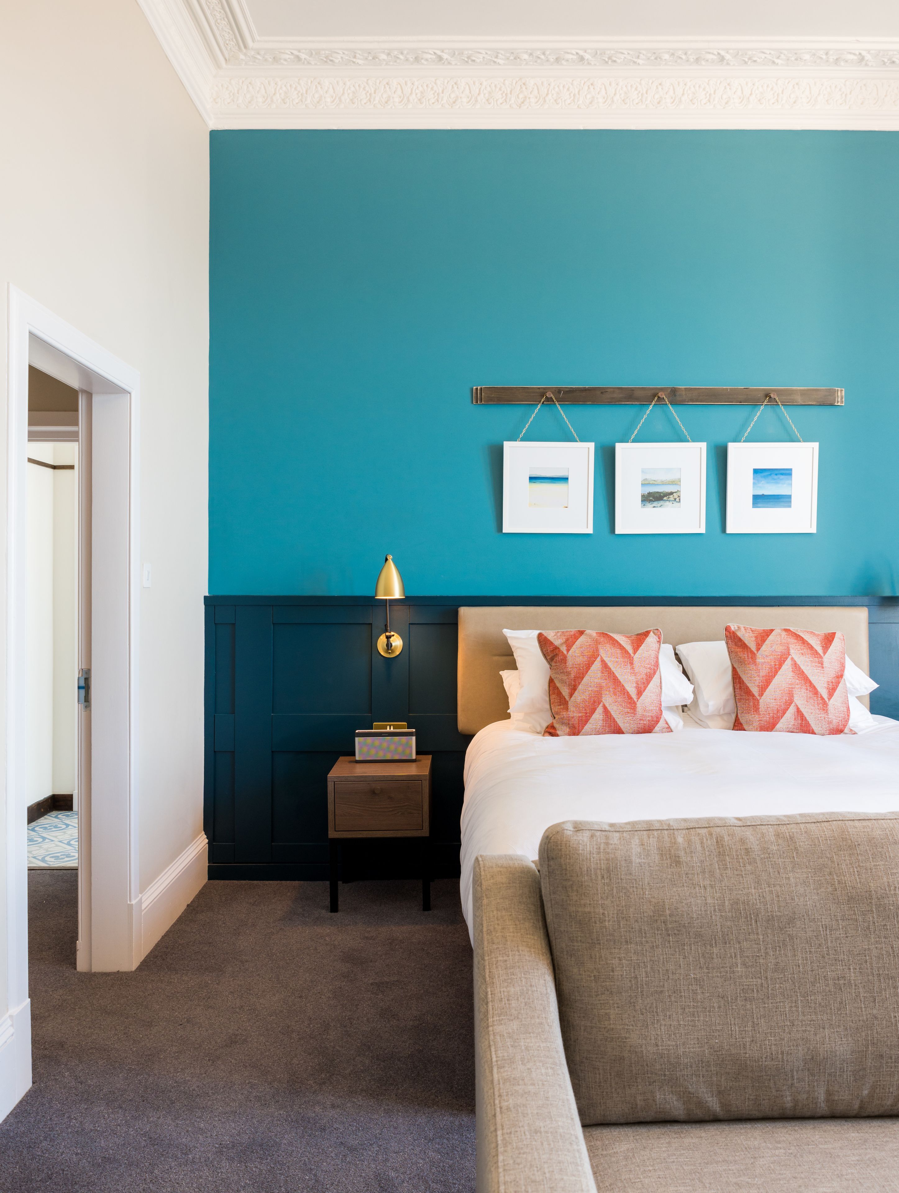 A bedroom with a teal wall, a bed with white and orange pillows, a nightstand with a lamp, and two framed pictures on the wall.