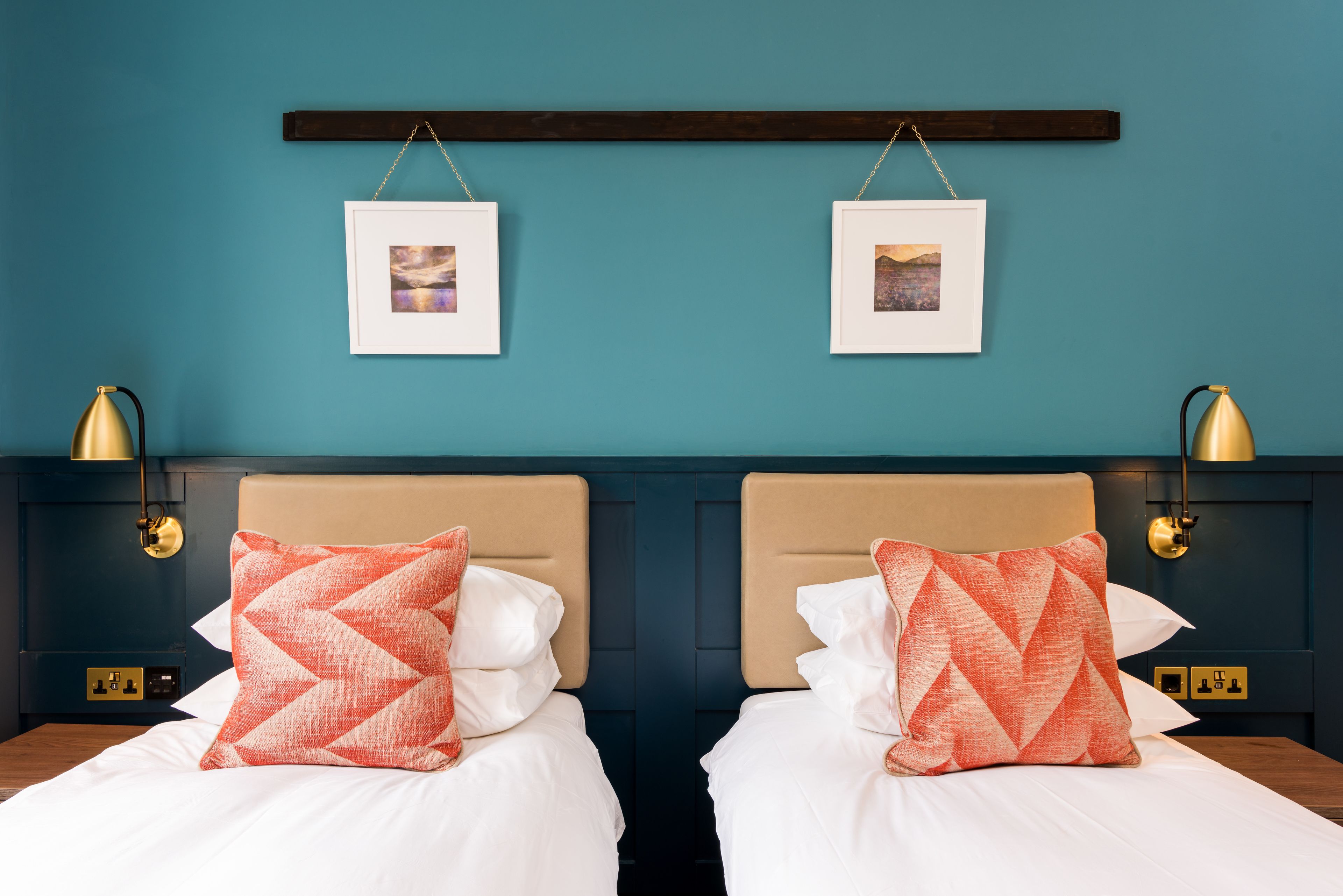 Deux lits jumeaux avec des têtes de lit beige, une literie blanche et des oreillers à motifs orange. Des tableaux encadrés sont accrochés au mur au-dessus des lits, et deux lampes murales sont fixées de chaque côté.