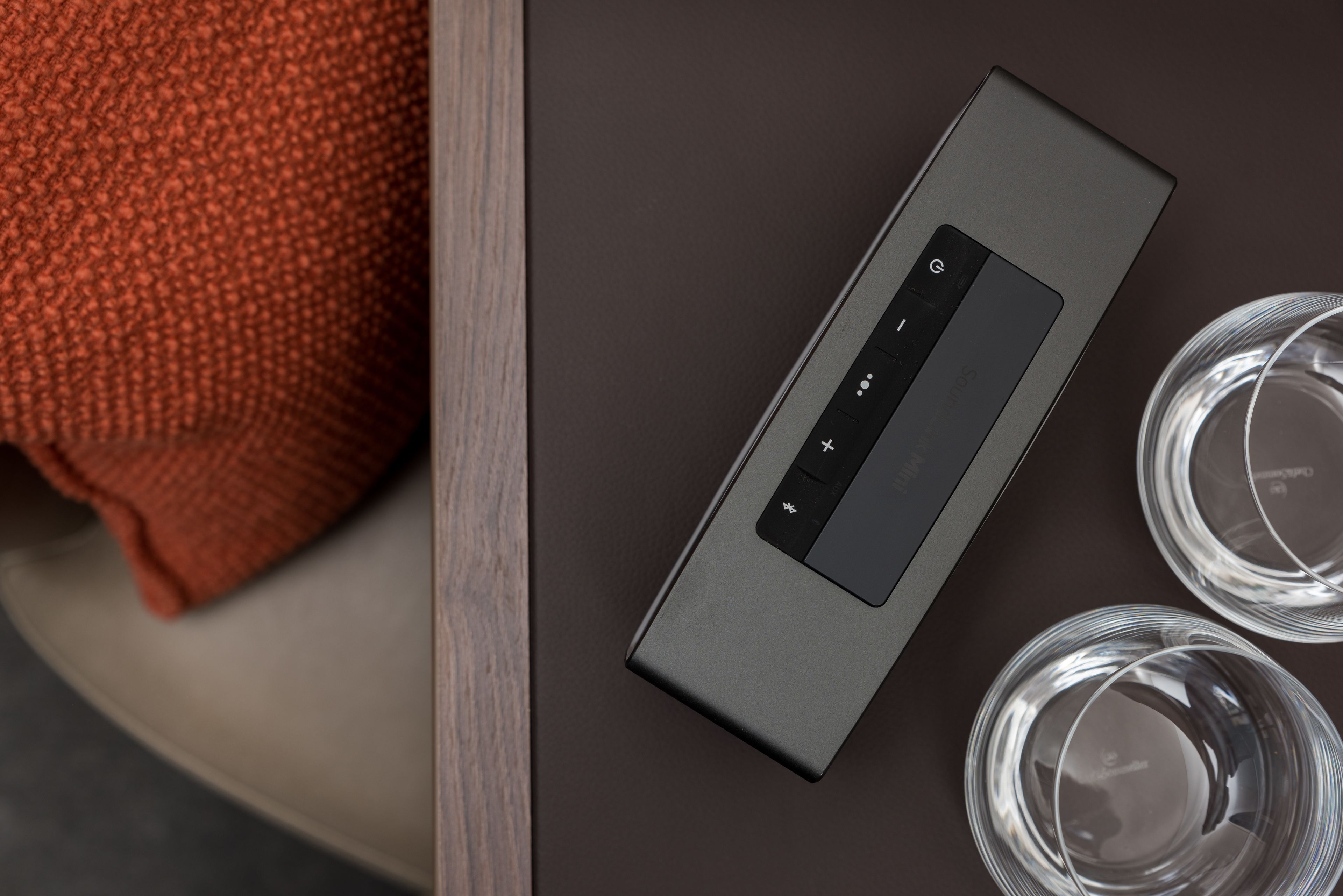 Una habitación moderna y minimalista con un dispositivo electrónico elegante y de color oscuro sobre una mesa, junto a un tejido tejido rojo y dos vasos transparentes.