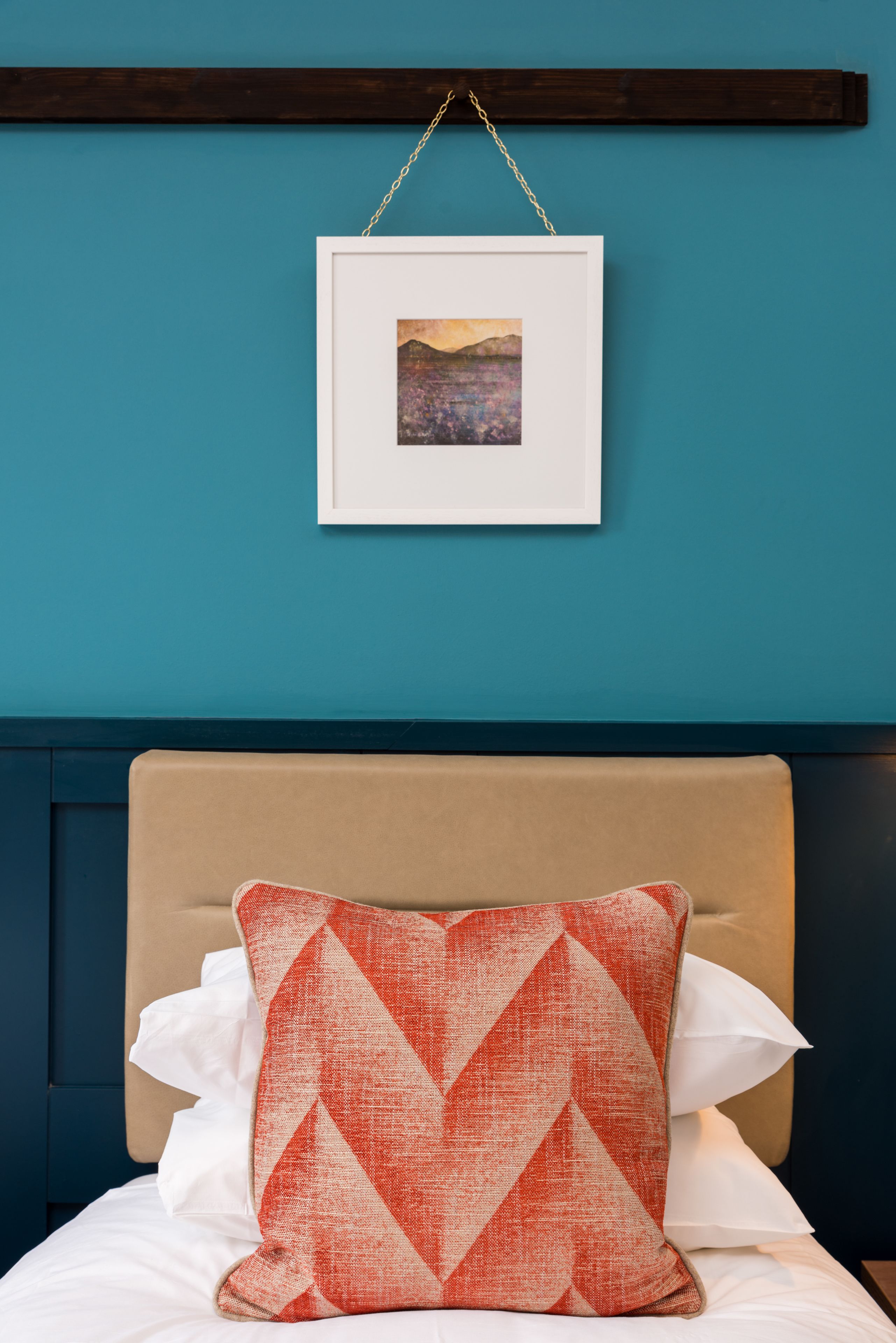 Una cama con cabecera color crema, almohadas blancas y una almohada con estampado rojo y blanco. Encima de la cama hay un cuadro en una pared azul.