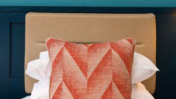 Una cama con cabecera color crema, almohadas blancas y una almohada con estampado rojo y blanco. Encima de la cama hay un cuadro en una pared azul.