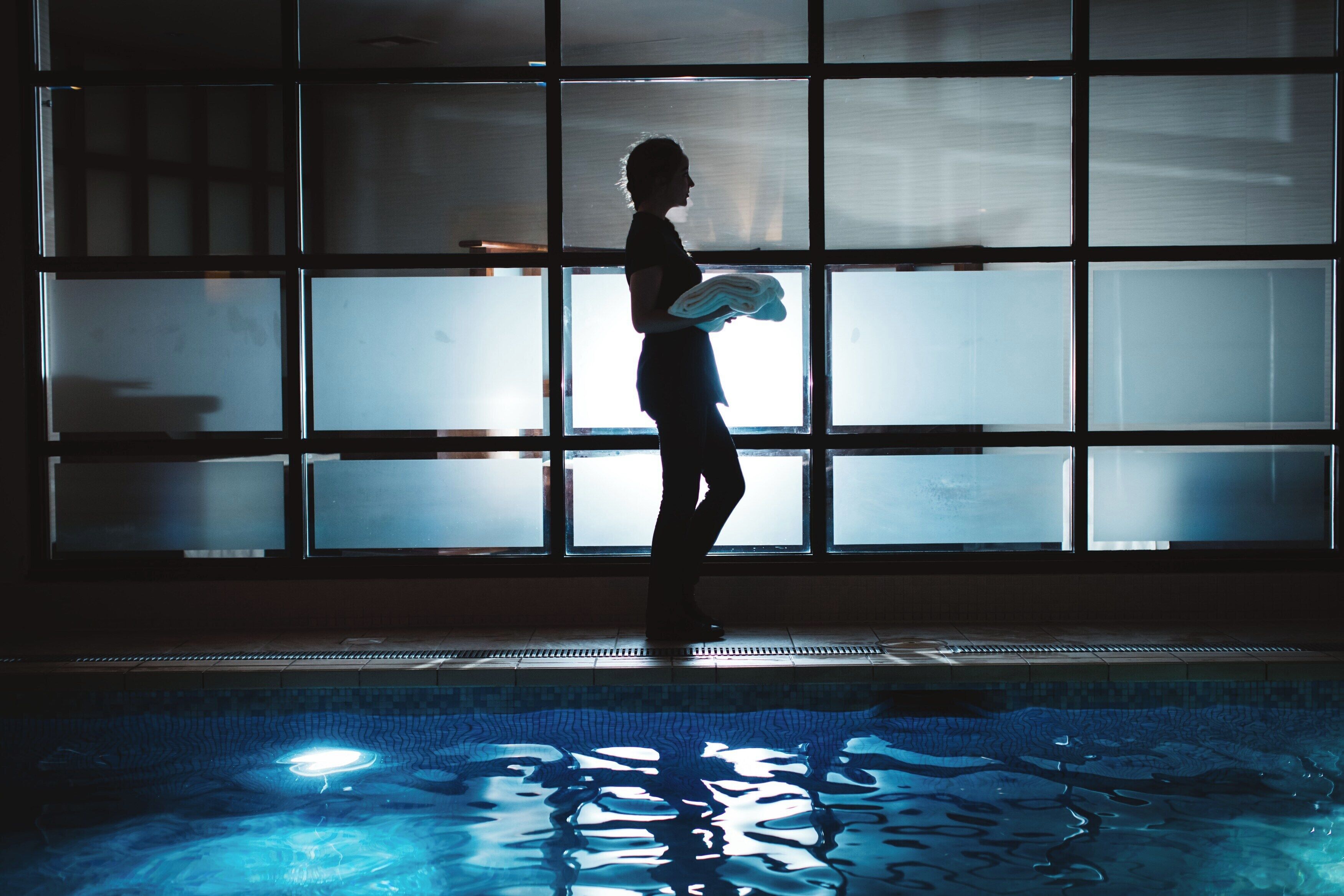 Una persona está de pie junto a una piscina con suelo de baldosas y una pared con ventanas al fondo.