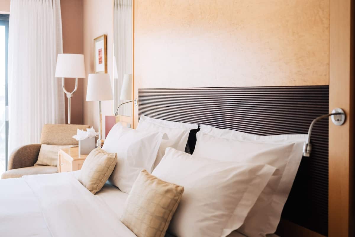 A neatly made bed with white linens and multiple pillows, a bedside table with a lamp, and a chair in a hotel room.
