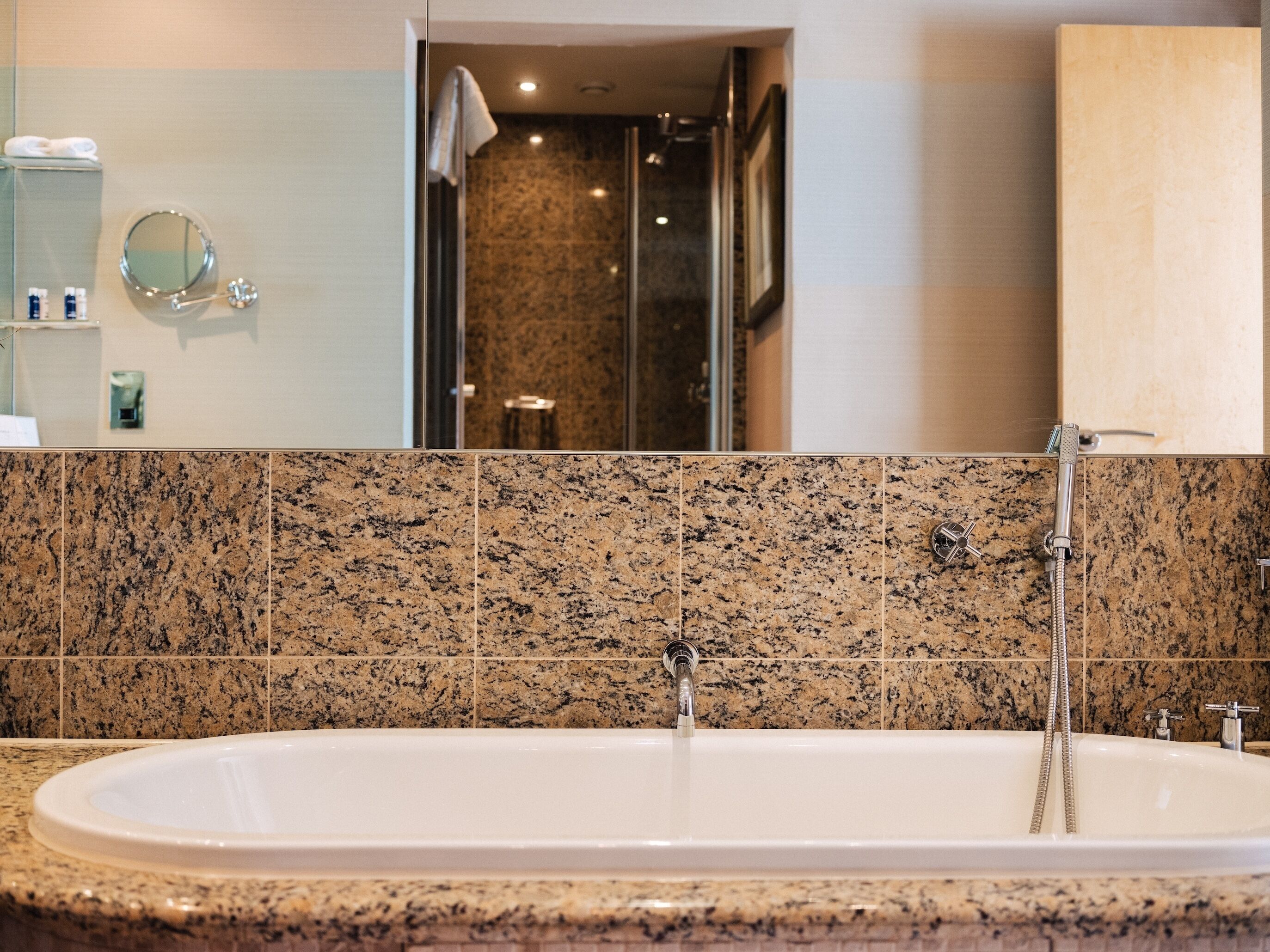 A bathroom with a large bathtub, granite tiles, a mirror, and a shower area.