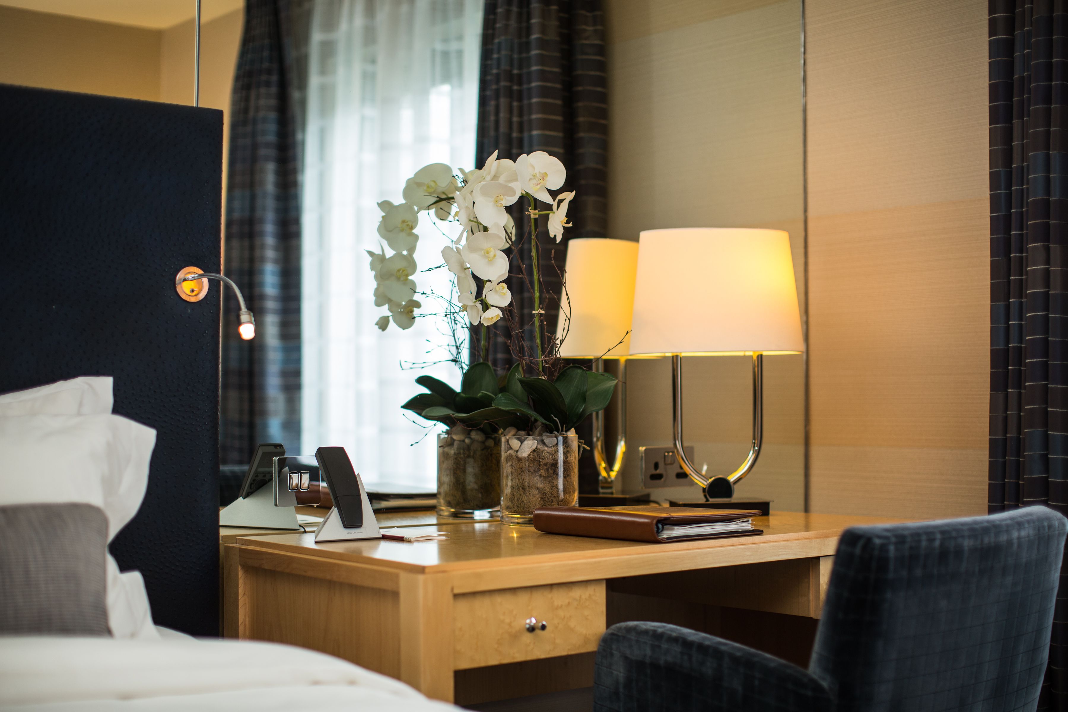 Une chambre d’hôtel avec un lit, un bureau doté d’une lampe, un téléphone et un vase contenant des fleurs blanches.